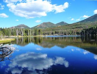Sprague Lake in Rocky Mountain National Park. Glassy blue lake with clouds reflected and above, surrounded by forest and mountain peaks in the background. A forest area the size of RMNP is cut down twice per year to create junk mail for US households.
