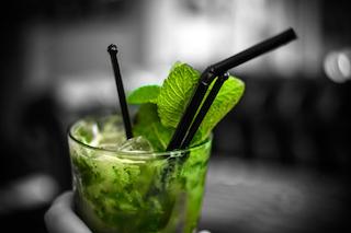 Mohito in glass with mint leaves, ice and black plastic straws, showing hand holding the glass at bottom of picture, with black and white background